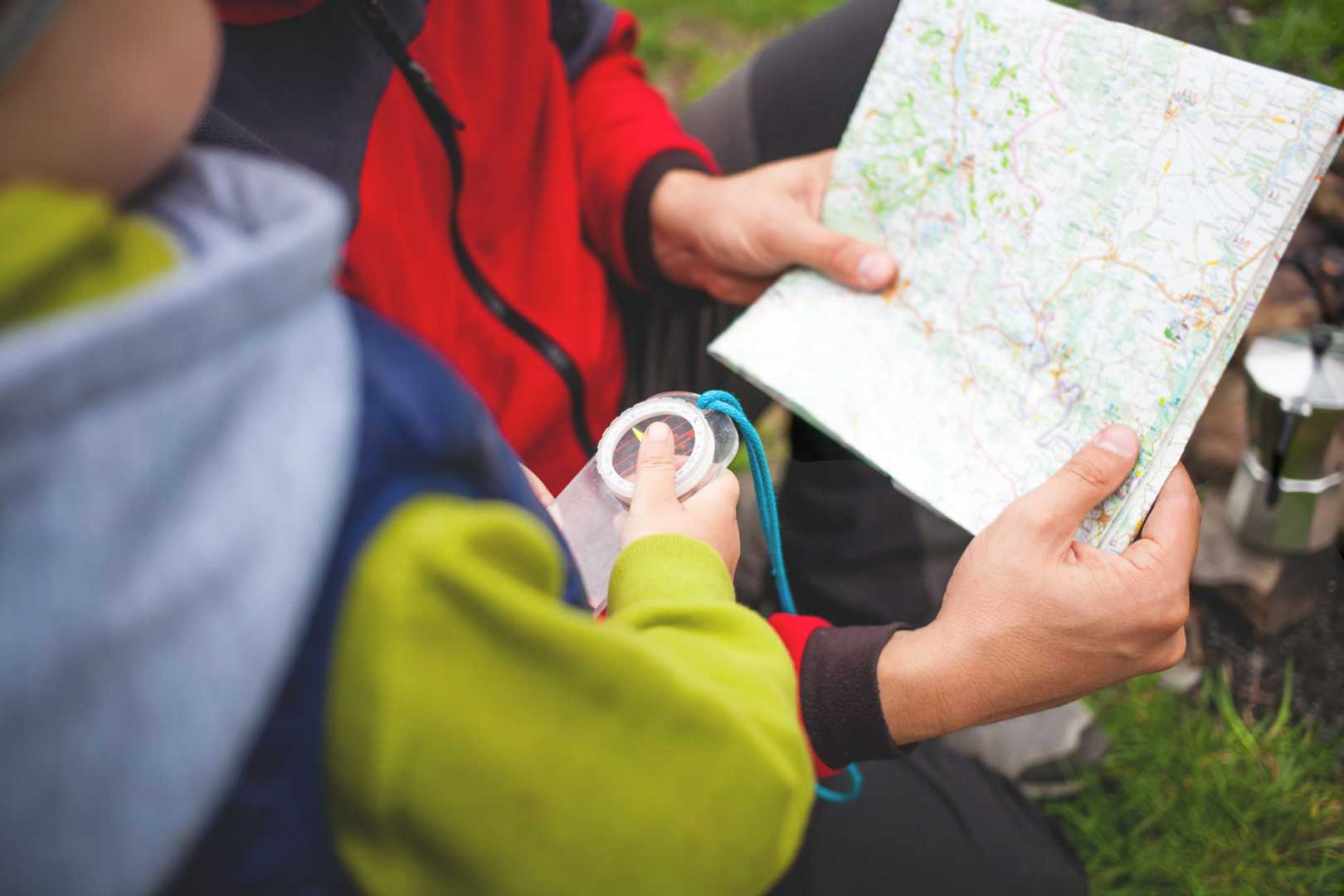 Kind beim Wandern mit Kompass in der Hand schaut auf eine Karte, die von einer anderen Person gehalten wird.