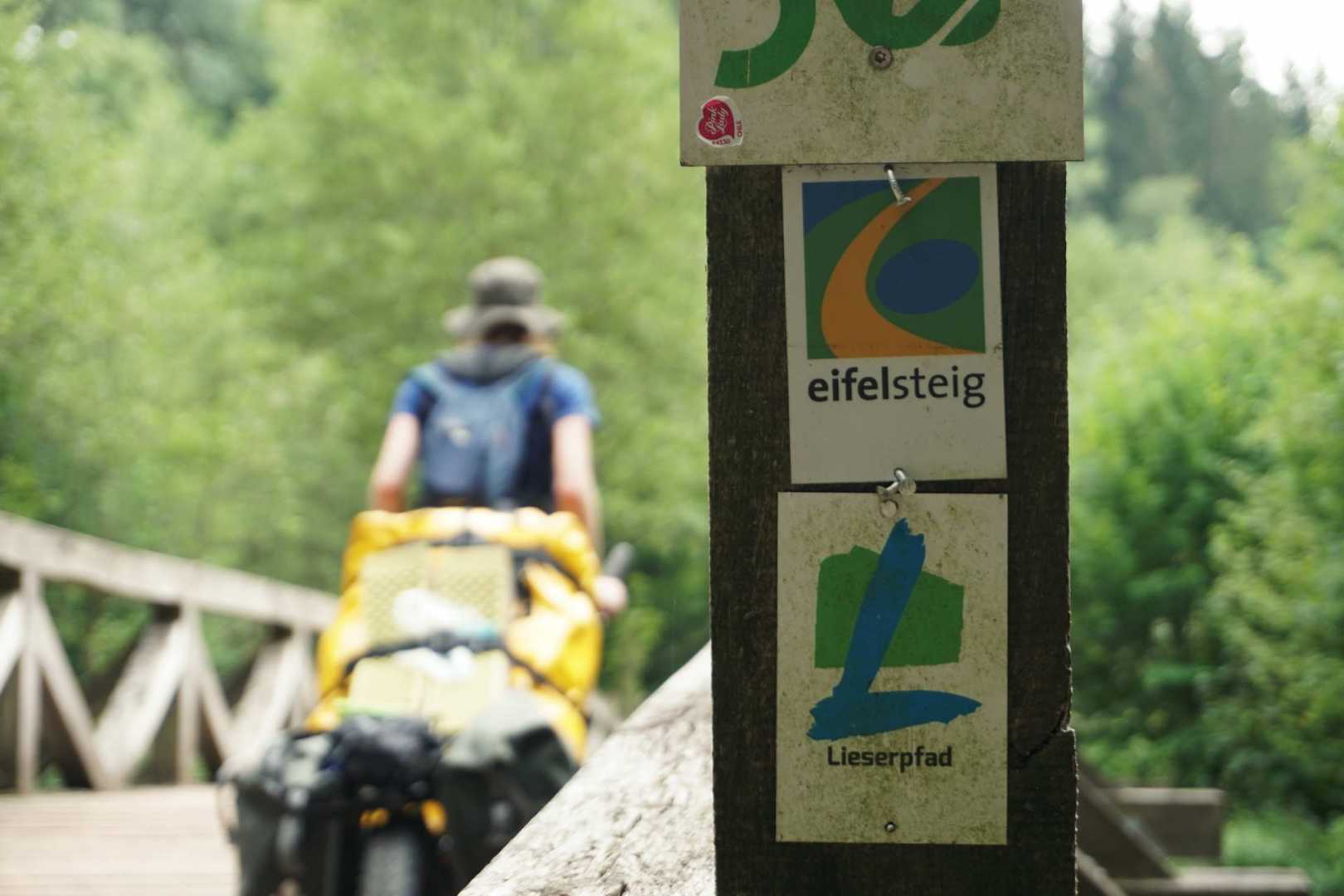 Jana fährt über eine Brücke mit dem Monowalker. Im Vordergrund markieren zwei Schilder den Eifelsteig und den Lieserpfad